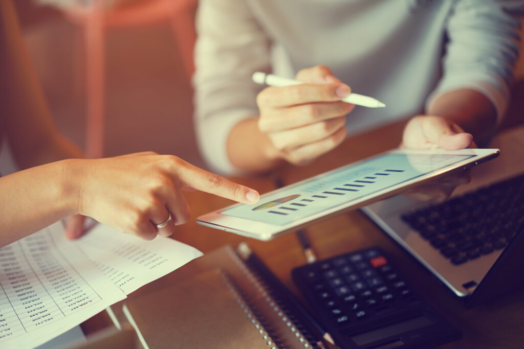 Two people analyse data on a tablet while reviewing documents and using a calculator, focused on charts and graphs.