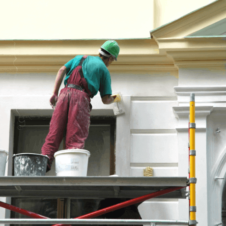 A worker in a green helmet applies paint to a building facade while standing on a scaffold, surrounded by buckets of paint representing soft FM services