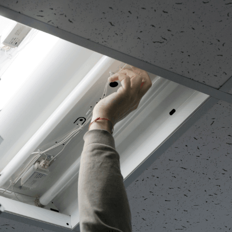 A hand reaching into a ceiling light fixture, adjusting wires and fluorescent bulbs in a white ceiling representing Hard Facilities Management services
